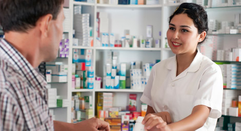 A pharmacist stood behind his counter with his arms folded staring out to the camera